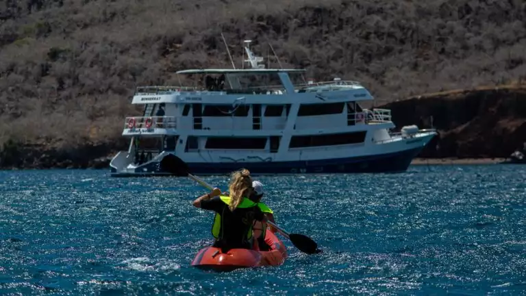 Tandem kayakers in red boat paddle calm blue sea toward a blue & white ship on a Monserrat Galapagos cruise.