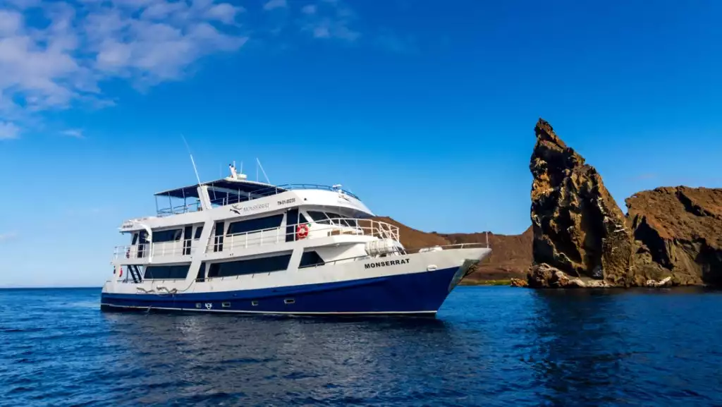 Monserrat boat in Galapagos with blue hull, white upper decks & 4 guest decks sitting beside Kicker Rock on a sunny day.