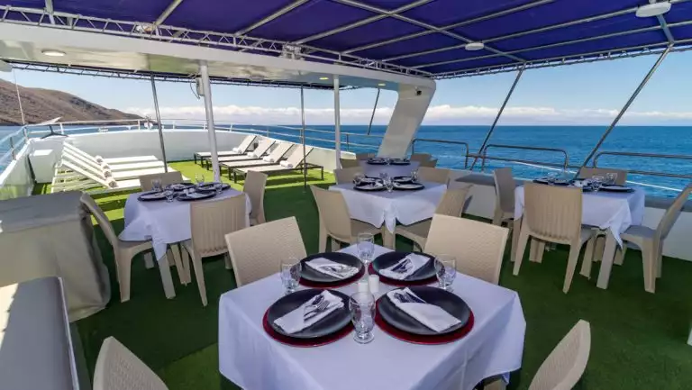 Tables set for outdoor dining under a blue awning near sun-soaked chaise loungers aboard Monserrat boat in Galapagos.