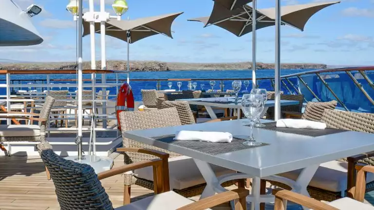 Outdoor dining area aboard Nat Geo Gemini. Square tables, chairs, umbrellas look out over blue Galapgos ocean and shoreline.