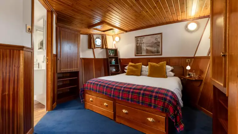 Scottish Highlander barge cabin with blue rug, wood paneled walls, portholes & wooden desk & chair.