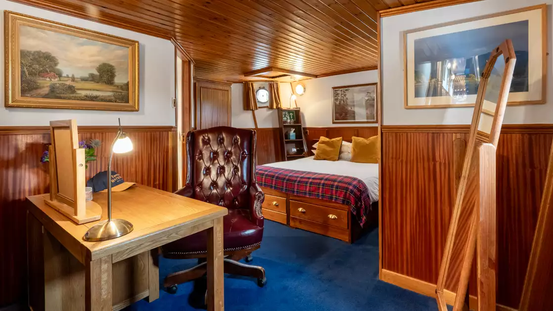 Scottish Highlander barge cabin with blue rug, wood paneled walls, portholes & wooden desk & chair.