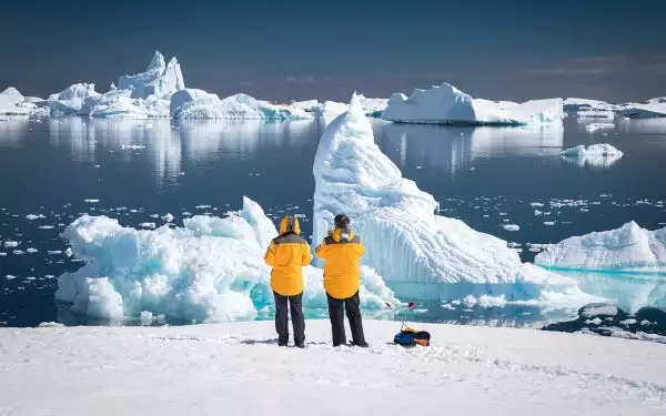 From a snowy shoreline two travelers in yellow parkas take in the polar landscape of snow and icebergs on an Antarctica trip