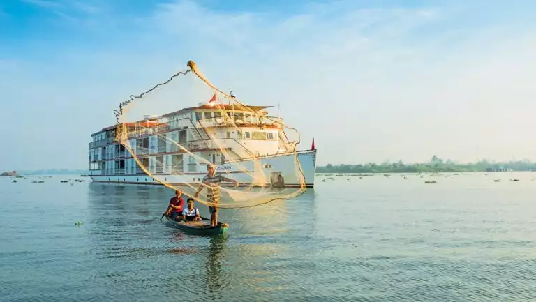 Jahan southeast asia cruise ship with white decks & red trim sits behind canoe of 3 fishermen casting a net on a sunny day,