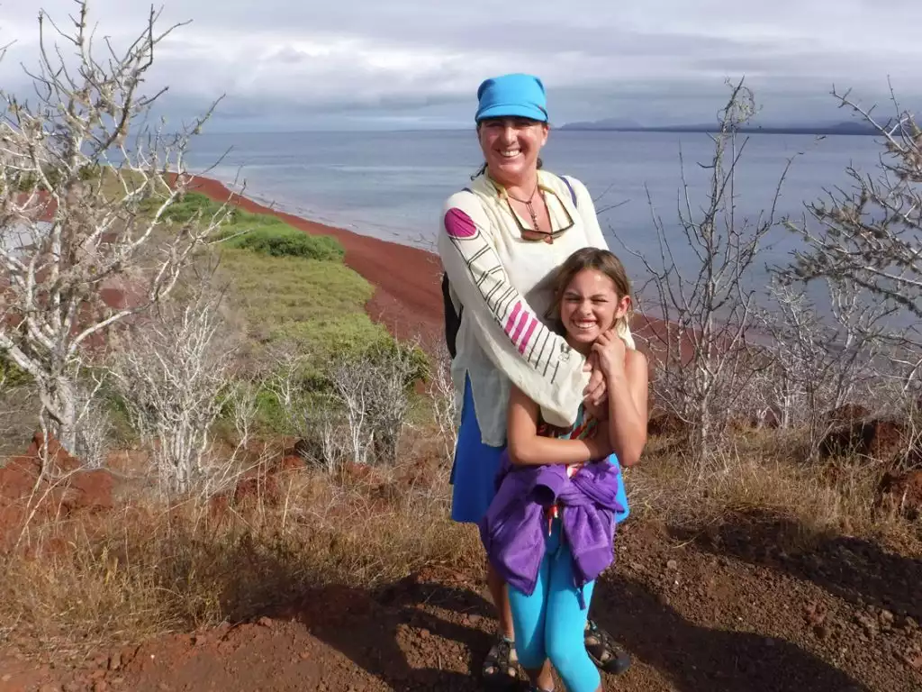 A mother in a blue hat stands hugging her daughter from behind on a hill in the Galapagos Islands