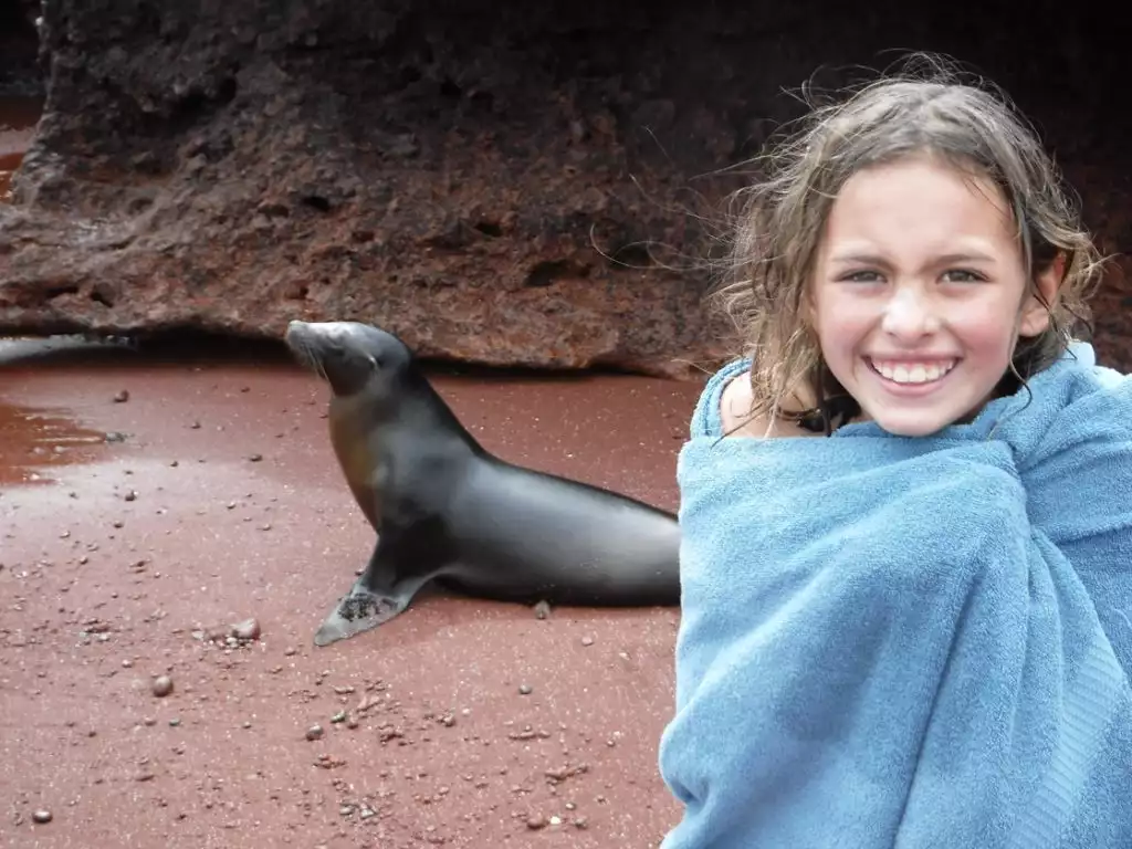 A young girl wrapped in a blue towel smiles in front of a Galapagos sea lion behind her