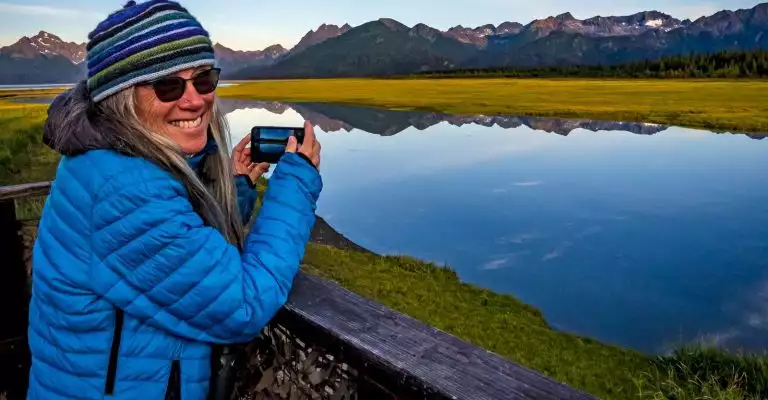 A traveler photographs the beautiful scenic mounatins and glass like waters with her iphone as she posses for her picture