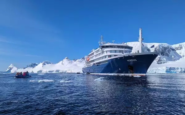 Ocean level view of a small Antarctica ship floating in blue water against a snowy icy background with a zodiac full of travelers cruising nearby.