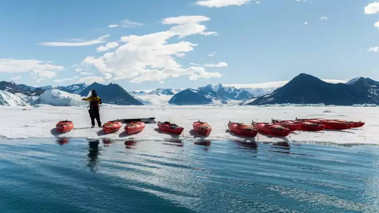 Small fleet of empty red kayaks lined up at water & ice's edge in the sun on the Commandant Charcot Unexplored Antarctica cruise.