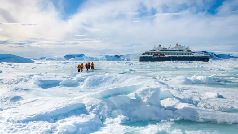 Small group of Unexplored Antarctica cruise guests in orange parkas walk over bumpy ice field near a small ship on a sunny day.