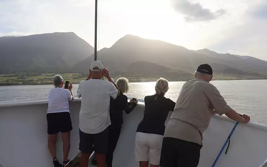 A group of passengers seen from behind on the bow of a ship looking at the Hawaiian Islands shoreline