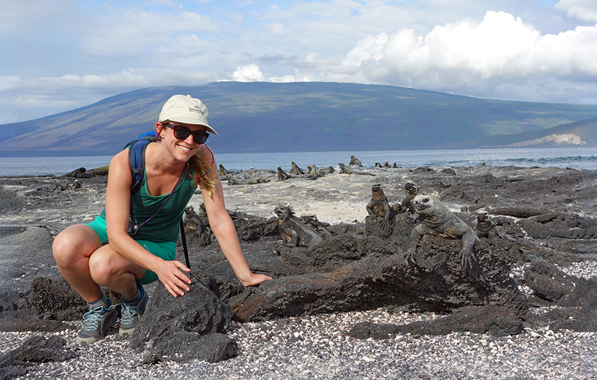 Woman in teal tank top & shorts kneels near gray marine iguanas sitting on sand & dark volcanic rock in the Galapagos Islands.