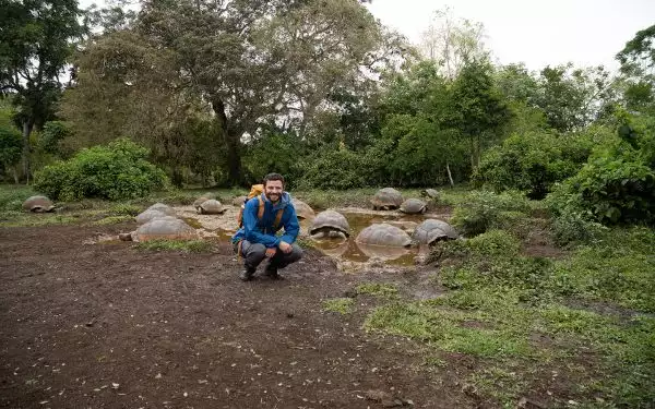 A man in a blue jacket and backpack squats in front of a small puddle full of Galapagos tortoises