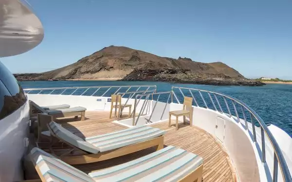The wood deck bow of a Galapagos yacht charter seen with striped sun lounge chairs and a barren island in front of it