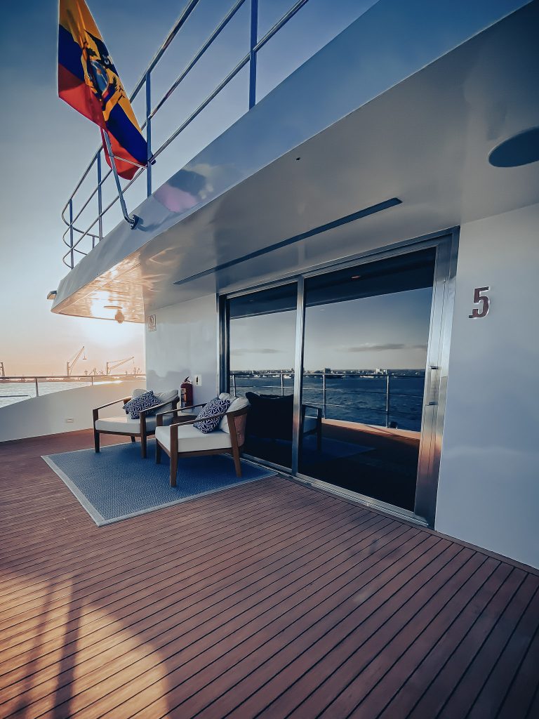 outdoor seating area aboard small cruise ship with 2 chairs a rug infront of large floor to ceiling windows as the sun sets