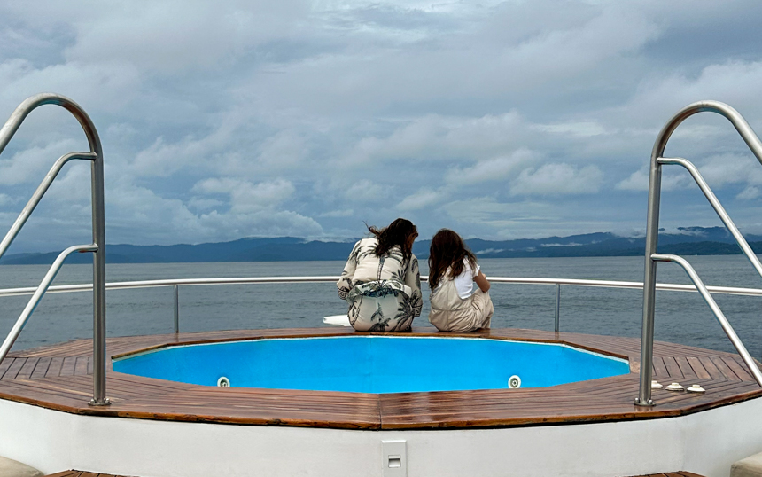 Mother and daughter sit on the end of a small cruise ship hot tub looking out at the stormy clouds