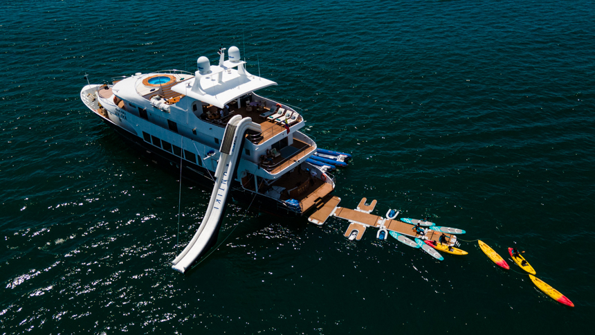 ariel view of small cruise ship with floating platforms to walk and swim on, paddleboards, a large slide and a swimmer around the stern of the boat