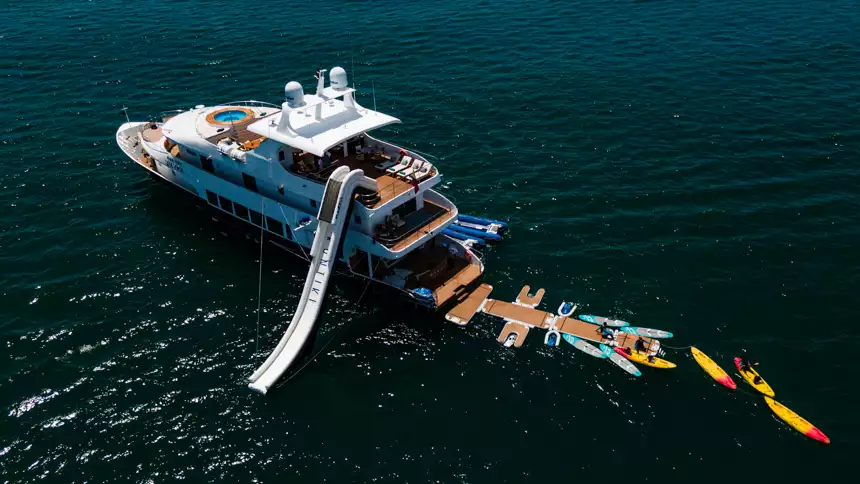 ariel view of small cruise ship with floating platforms to walk and swim on, paddleboards, a large slide and a swimmer around the stern of the boat 