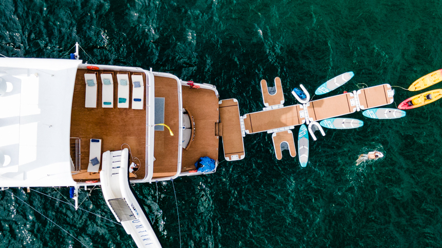 ariel view of small cruise ship with floating platforms to walk and swim on, paddleboards, a large slide and a swimmer around the stern of the boat