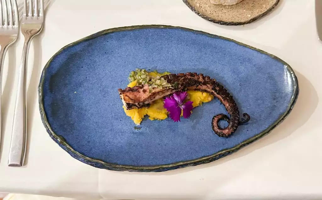 octopus beautifully plated over yellow vegetable on a blue plate with a purple flower for some color