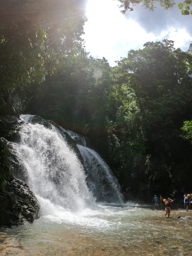Waterfall flows down the side of massive rocks in a green jungle as travelers cool off in the water below