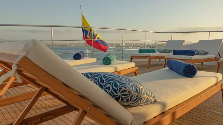 Wood chaise loungers with white pads & blue throw pillows on teak deck of Kontiki yacht at sunset in Costa Rica.