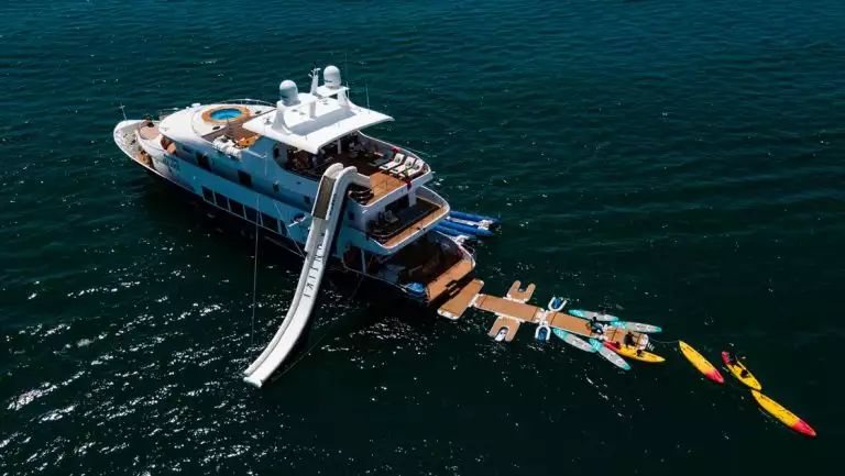 Aerial view of Kontiki yacht with blue hull, white & teak upper decks, waterslide, floating aft dock, paddleboards & kayaks.