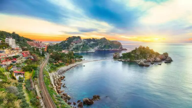 Aerial view of Taormina, Sicily at sunset with rocky, green islands & rail line along idyllic bays, seen on a cruise to Malta.