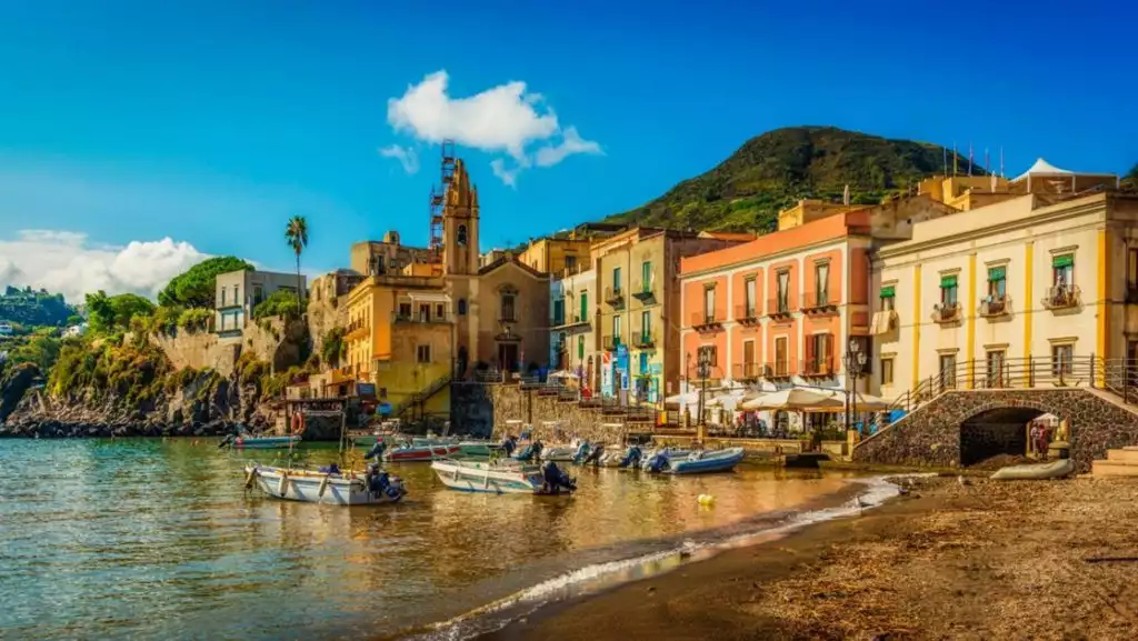 Lipari, Italy with sandy beach, calm water, fishing boats & various colorful buildings under a blue sky.