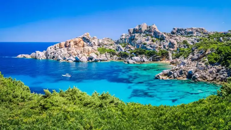 swimming cove off the ocean with lush green bushes and large rocks surrounding a small skiff in bright blue waters