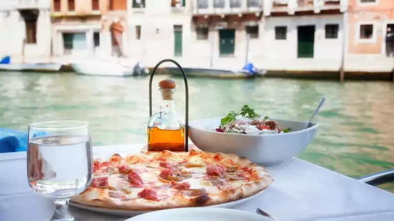 A pizza plated on a white linen table with side salad and a glass of water overlooking a canal