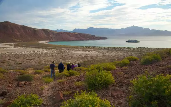 In Baja, travelers walk towards a teal colored bay with a single small ship anchored off shore surrounded by red desert mountains