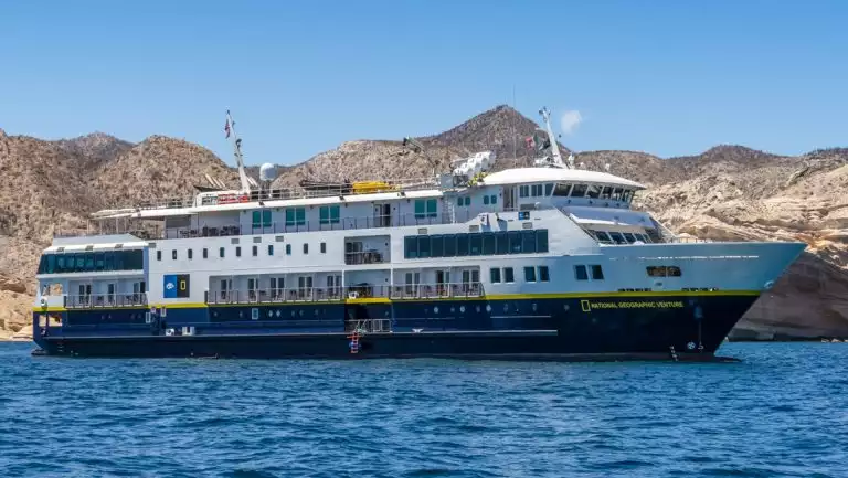 National Geographic Venture small ship with blue hull & white upper decks sits offshore of arid beige mountains in Baja.