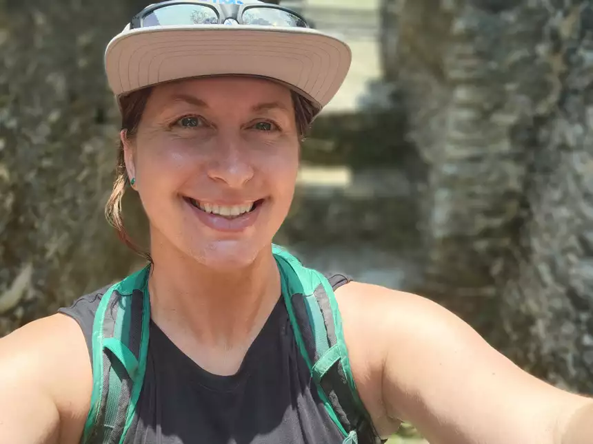 A woman in a hat and green backpack smiles for a selfie in front of trees