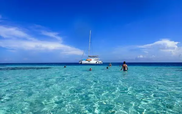 Family of travelers on a Belize small ship cruise swim in sparkling turquoise & blue sea by a small white catamaran in the sun.