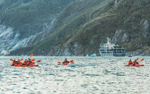 Tandem kayakers in red boats paddle choppy water near small dark blue ship & rocky, vegetated cliffs on a Chile cruise.