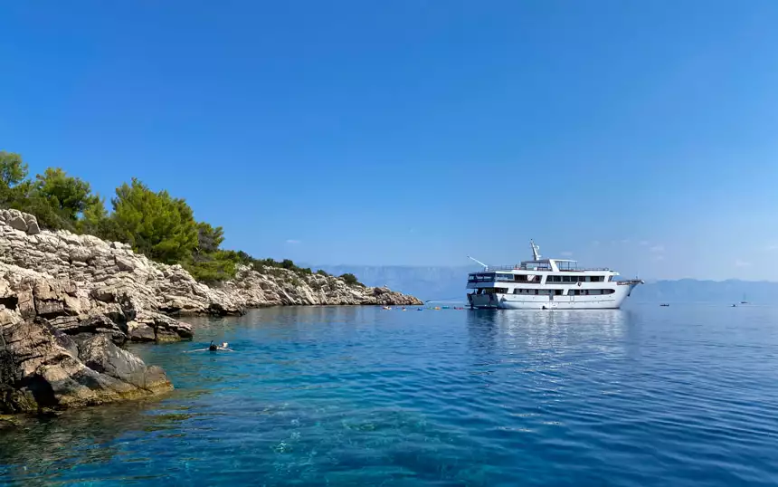 A Croatia small ship cruise yacht seen at anchor in a swim stop with a person snorkeling in the water and others swimming behind the ship