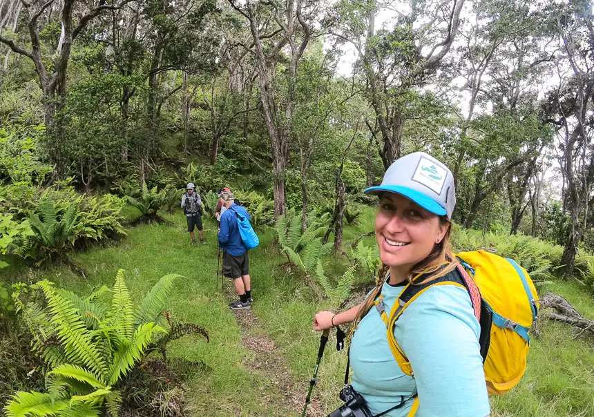 A female traveler in a hat stands on a lush tropical trail looking back smiling at the camera