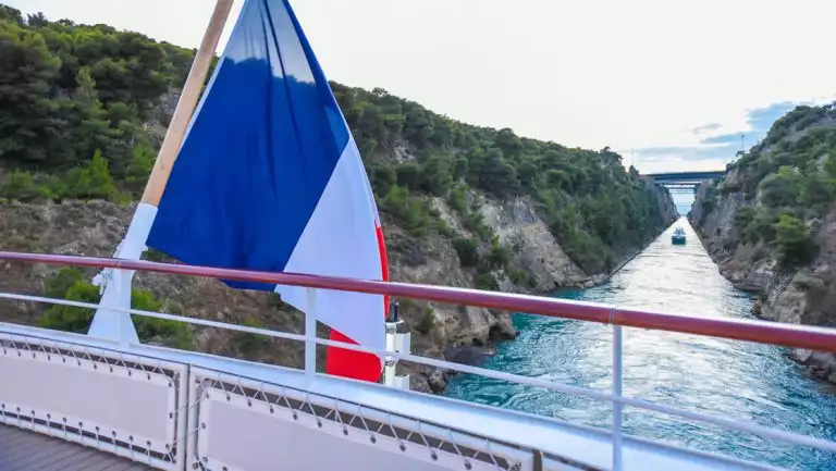 Aft of modern small ship with French flag as it sails the tight Corinth Canal with blue sea & green cliffs on either side.