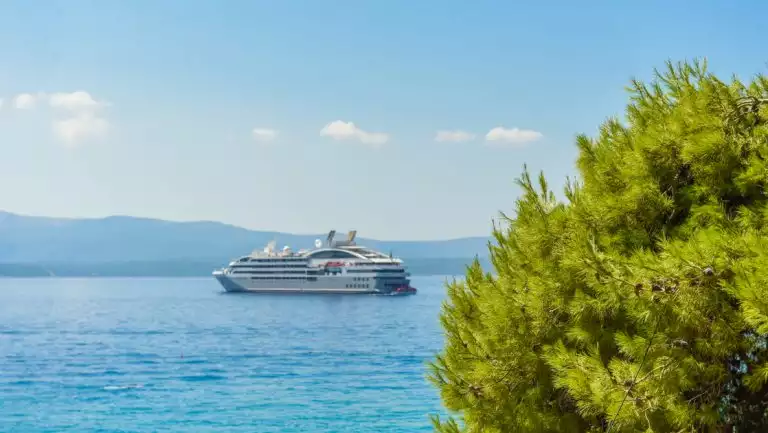 Small gray expedition ship sits in teal sea offshore from a bright green tree during a Dalmatian Coast & Ionian Sea cruise.