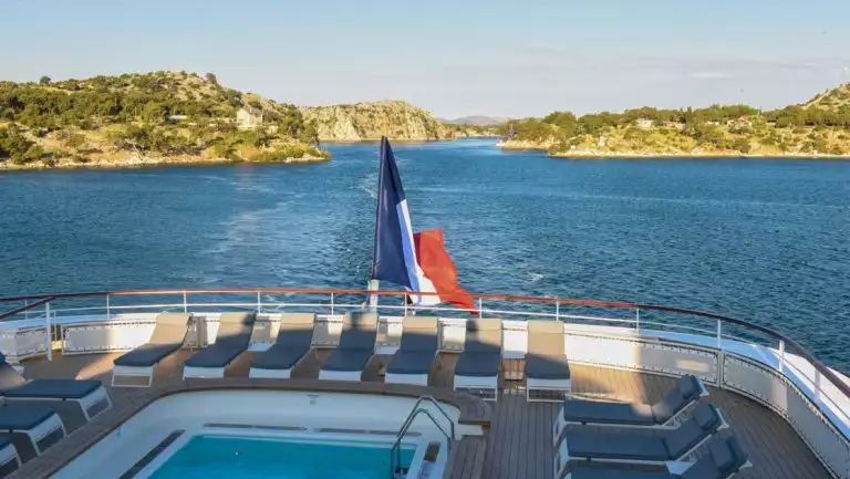 Aft of small ship with French flag & pool as it cruises among green & gold hillsides on a Dalmatian Coast & Ionian Sea cruise,