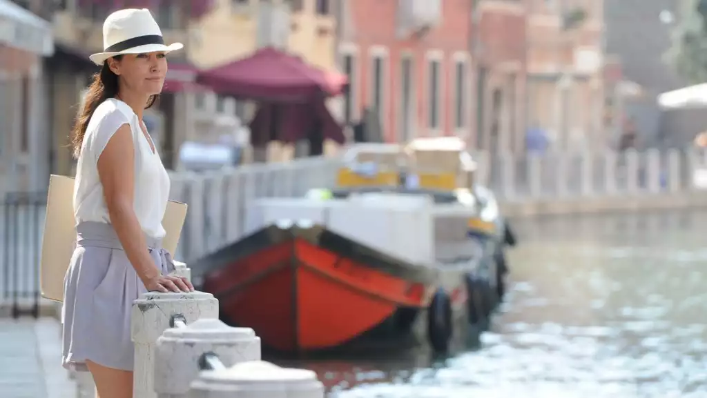 Woman in white hat & blouse & gray shorts stands along Venice's Grand Canal with a small riverboat nearby & colorful buildings.