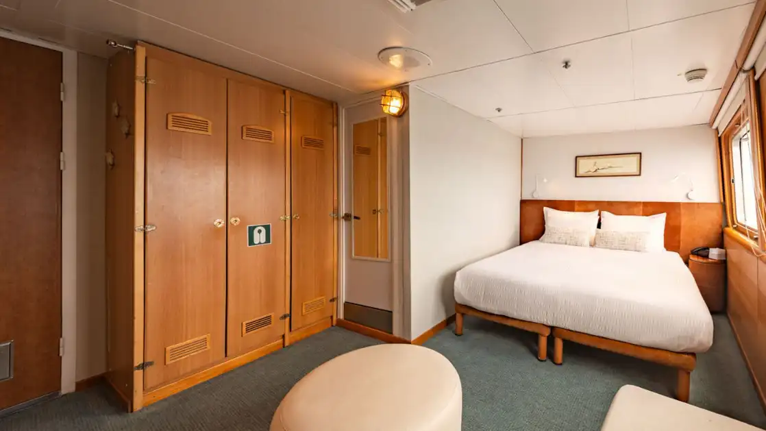 Legend Suite on Galapagos Legend cruise ship with double bed in white, wood accents & wardrobe, ottoman & view window.