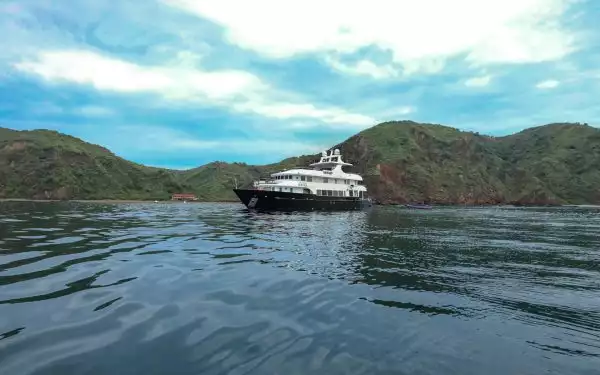 Small kontiki yacht floats in a clam by on a cloudy day in front of beautiful green rolling hills