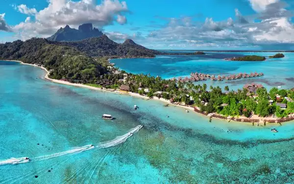 An aerial view of a small strip of land in Bora Bora with a small ship sailing in the teal water and thatch roof huts on the other side of the island