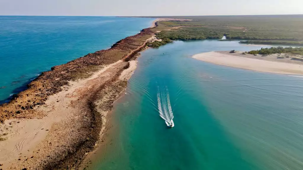 Small white tender vessel cruises an inlet of teal sea by white-sand beach on the Paspaley Pearl Pearling Coast cruise.