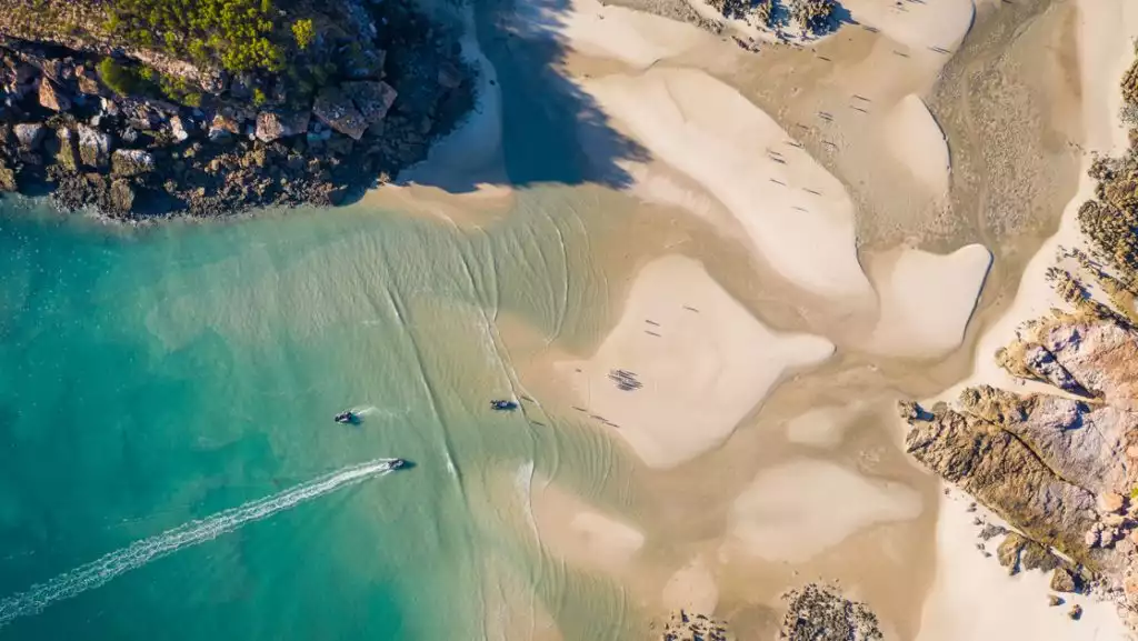 Aerial view of Zodiac boats cruising to & from white-sand beach beside chunky rock cliffs in Australia's Kimberley region.