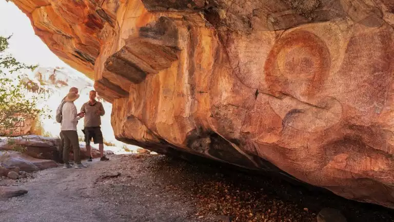 Guide & 2 PASPALEY PEARL Unexplored Kimberley travelers stand beside red cliff with ancient carvings in Australia.