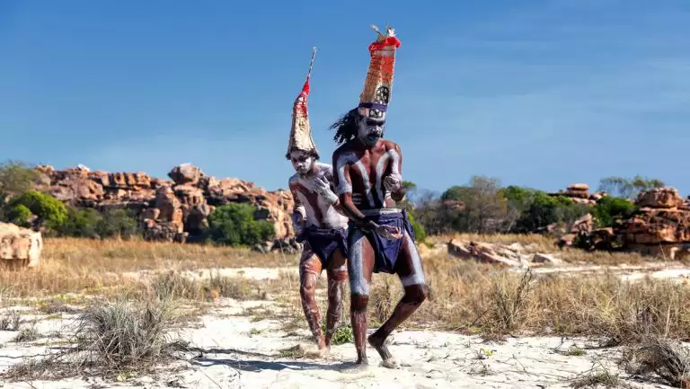 2 indigenous Australians wear tall, bright hats & white face paint while walking through sand dunes in the Kimberley.