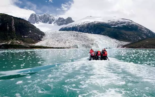 Zodiac boat with Patagonia cruise guests in orange life jackets motors toward Pia Glacier with jagged peaks behind in the sun.